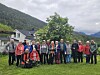 En gruppe står samlet på en gresslette med hus, trær og fjell i bakgrunnen.