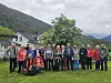 En gruppe står samlet på en gresslette med hus, trær og fjell i bakgrunnen.