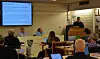 Foredragsholder står ved talerstol i et bibliotekrom mens publikum sitter og følger en presentasjon på skjerm.