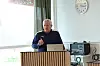 Mann ved talerstol med mikrofon og laptop i et lyst rom med klokke på veggen.