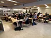 Voksne sitter ved små bord i et lyst møtelokale med veggopphengte bilder.
