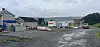 Gruslagt industriområde med lagerbygg, containere og lastebiler ved en fjord under grå himmel.
