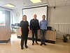 Tre personer står foran et lerret i et møterom med vinduer og bord langs veggen.