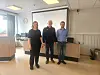 Tre personer står foran et lerret i et møterom med vinduer og bord langs veggen.