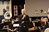 Dirigent står foran et orkester og holder mikrofon på en scene innendørs.