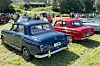 To klassiske veteranbiler, en blå og en rød, står parkert på en grasbakke med folk og flere biler i bakgrunnen.