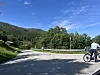 To syklister på en landevei ved foten av grønne åser og skog under blå himmel.