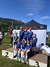 Barn og unge i blå drakter står og sitter på en seierspall foran et klubbelt i fjellandskap.