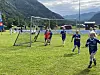 Barn i fotballdrakter løper på en gressbane med mål, fjell og hus i bakgrunnen.