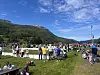 Folk står og går på en gresslette med fjell, flaggstang og telt i bakgrunnen.