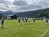 Barn i blå drakter spiller fotball på en gressbane med fjell i bakgrunnen.