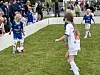 Barn spiller fotball på en gressbane mens tilskuere står langs sidelinjen bak gjerder.