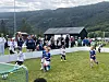 Barn spiller fotball på en gressbane med publikum langs sidelinjen og skogkledde åser i bakgrunnen.