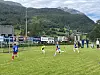 Fotballspillere på en gressbane med mål og fjell i bakgrunnen.