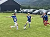 Barn i blå og røde fotballdrakter spiller på en gressbane med biler og fjell i bakgrunnen.
