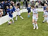 Barn i fotballdrakter kjemper om ballen på en gressbane med publikum i bakgrunnen.