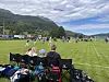 Fotballspillere og tilskuere på en gressbane med fjell og skyet himmel i bakgrunnen.