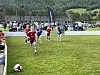 Barn spiller fotball på en gressbane med publikum og skogkledde åser i bakgrunnen.