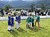 Barn i blå og grønne fotballdrakter står på en gressbane med tilskuere og fjell i bakgrunnen.