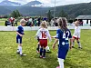 Barn i fotballdrakter spiller på en gressbane med fjell og tilskuere i bakgrunnen.
