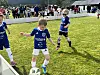 Barn i blå fotballdrakter spiller på en gressbane med publikum i bakgrunnen.