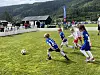 Barn spiller fotball på en grønn bane med publikum og fjell i bakgrunnen.