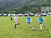 Tre barn spiller fotball på en gressbane med biler og skog i bakgrunnen.