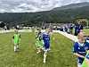 Barn i fotballdrakter løper på en gressbane med publikum og fjell i bakgrunnen.