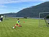 Ung fotballspiller skyter på mål på grønn bane med fjell, vann og mål i bakgrunnen.