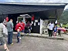 Folk står i kø utenfor et serveringssted med skrått tak og utsikt mot fjell i bakgrunnen.