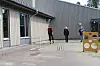 Tre personer står på en skoleplass med små hinder foran en grå bygning.