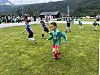 Barn spiller fotball på en gressbane med fjell og publikum i bakgrunnen.