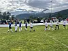 Ungdomsspillere på en grønn fotballbane med tilskuere og fjell i bakgrunnen.