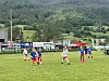 Fotballspillere i rødt og blått på en gressbane med skog og åser i bakgrunnen.