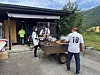 Tre personer står ved et bord med mat og grillutstyr utenfor en tømmerbygning i fjellandskap.