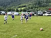 Barn løper på en fotballbane mot en ball, med biler og skog i bakgrunnen.