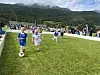 Barn spiller fotball på en gressbane med tilskuere og fjell i bakgrunnen.