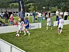Barn spiller fotball på en liten gressbane med publikum langs sidelinjen.