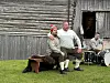 To menn sitter på en benk foran et gammelt trebygg, mens en trommeslager sitter ved et trommesett i bakgrunnen.
