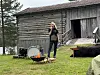 En kvinne står på gress foran en gammel trebygning med utstyr og en liten bålplass i forgrunnen.