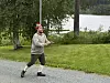 En mann i rød lue går på en grusvei ved en innsjø med trær og gress rundt.