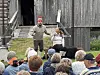 To skuespillere opptrer foran en sittende publikumsmengde ved en gammel trebygning utendørs.