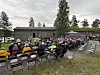 Et stort publikum sitter på stoler ute ved en dam, med en låve og trær i bakgrunnen.