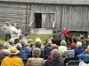 Skuespillere opptrer foran et tregammelt bygning ved et utendørs arrangement, mens publikum sitter samlet på stoler og benker i regnvær.