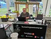 En mann som sitter bak et informasjonsbord med laptop, brosjyrer og skilt i et lyst telt.