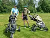 To personer står på en golfbane med traller og golfutstyr foran trær og gress.
