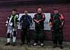 Fire personer i skytterklær står ved en rød vegg foran et delvis synlig skilt.