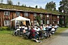 Mange personer sitter i stoler foran ei gammel trønderlån med hvit paviljong på tunet.