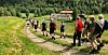 Ei gruppe vandrere går på en smal sti gjennom grønt fjellandskap med skog og en bygning i bakgrunnen.