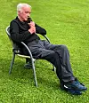 En eldre mann sitter i stol på gress og holder snakker i mikrofon.